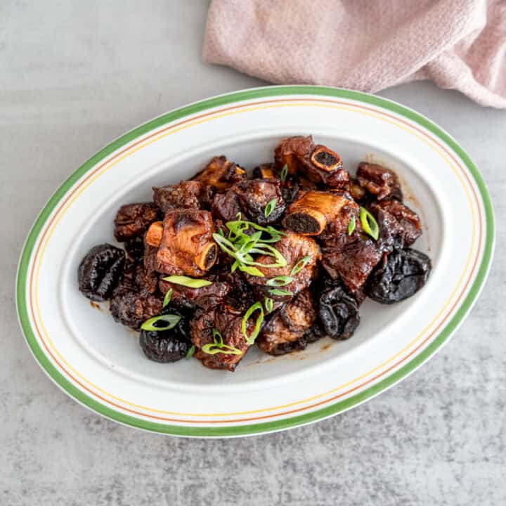 Close-up of tender Chinese braised spareribs with prunes (酸梅排骨) in a rich, glossy sauce, garnished with green onions, served in a white ceramic dish.