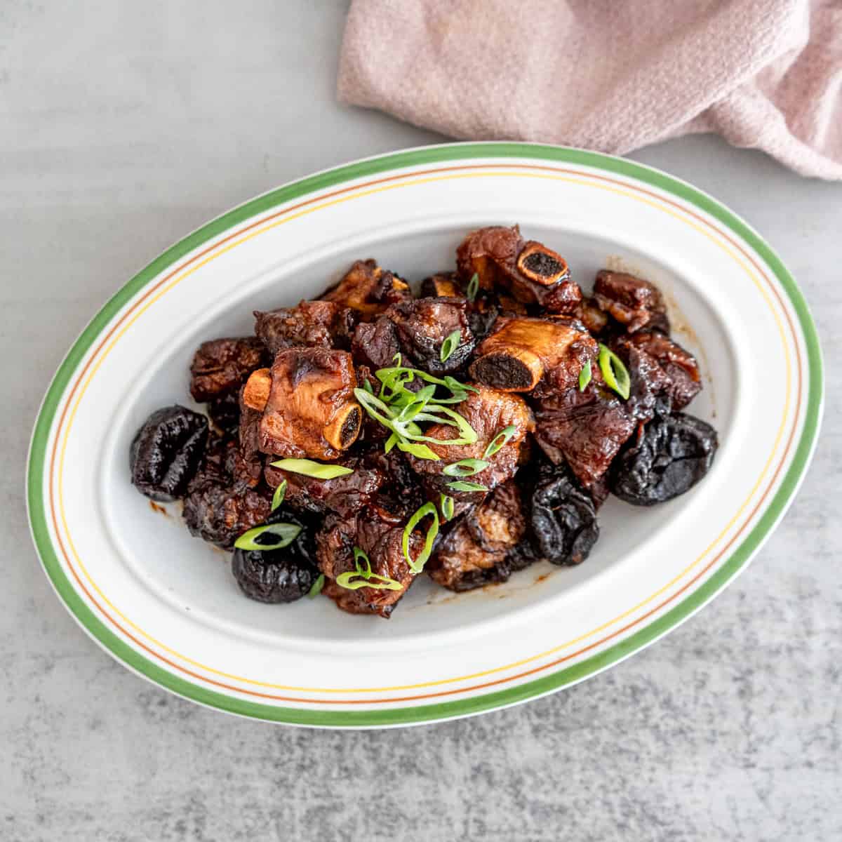 Close-up of tender Chinese braised spareribs with prunes (酸梅排骨) in a rich, glossy sauce, garnished with green onions, served in a white ceramic dish.