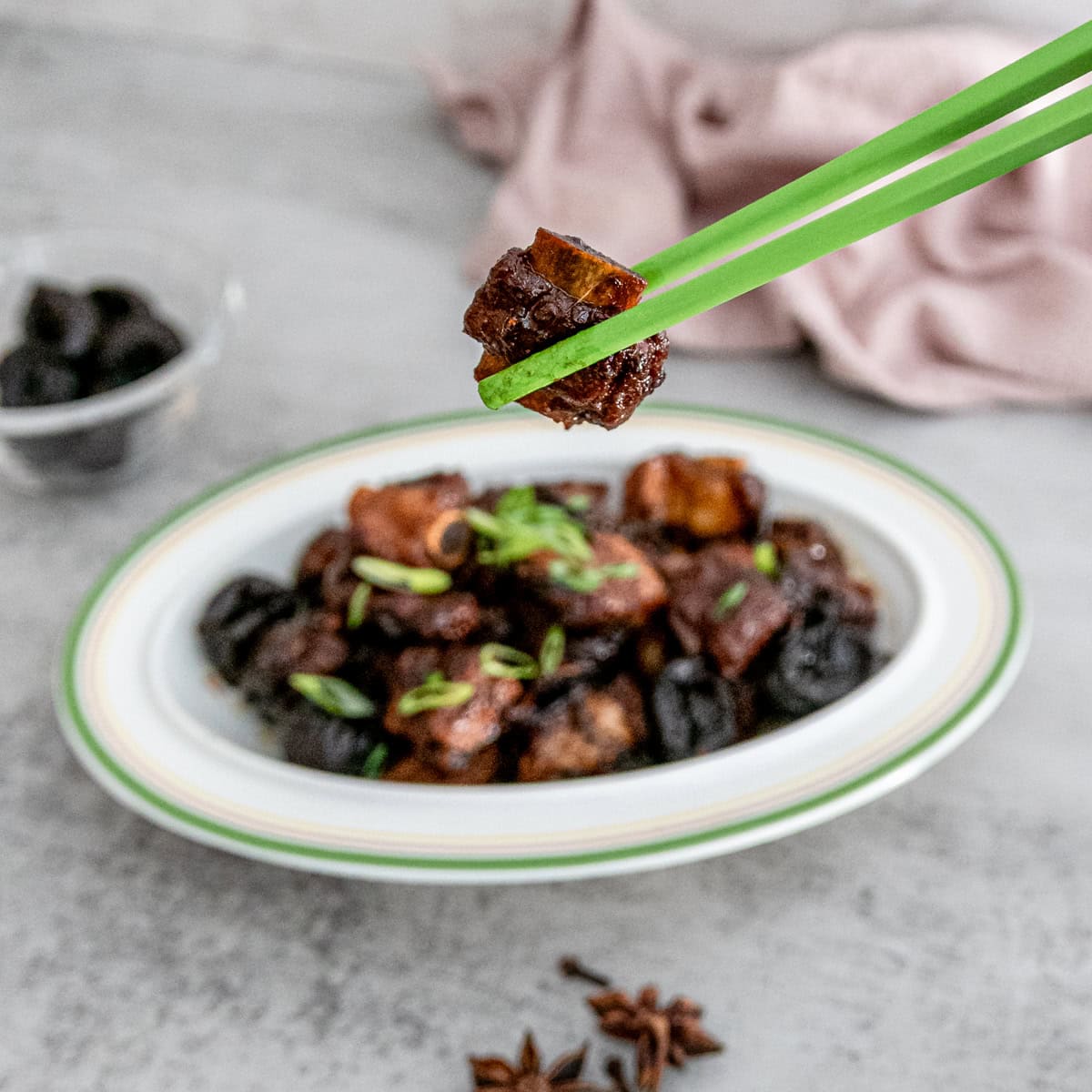 Close-up of tender Chinese braised spareribs with prunes (酸梅排骨) in a rich, glossy sauce, garnished with green onions, served in a white ceramic dish.