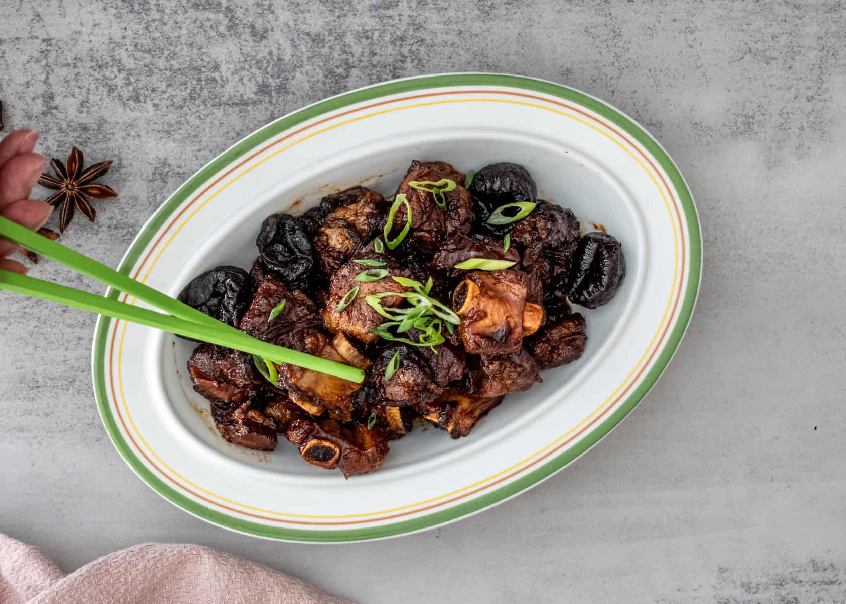 Close-up of tender Chinese braised spareribs with prunes (酸梅排骨) in a rich, glossy sauce, garnished with green onions, served in a white ceramic dish.