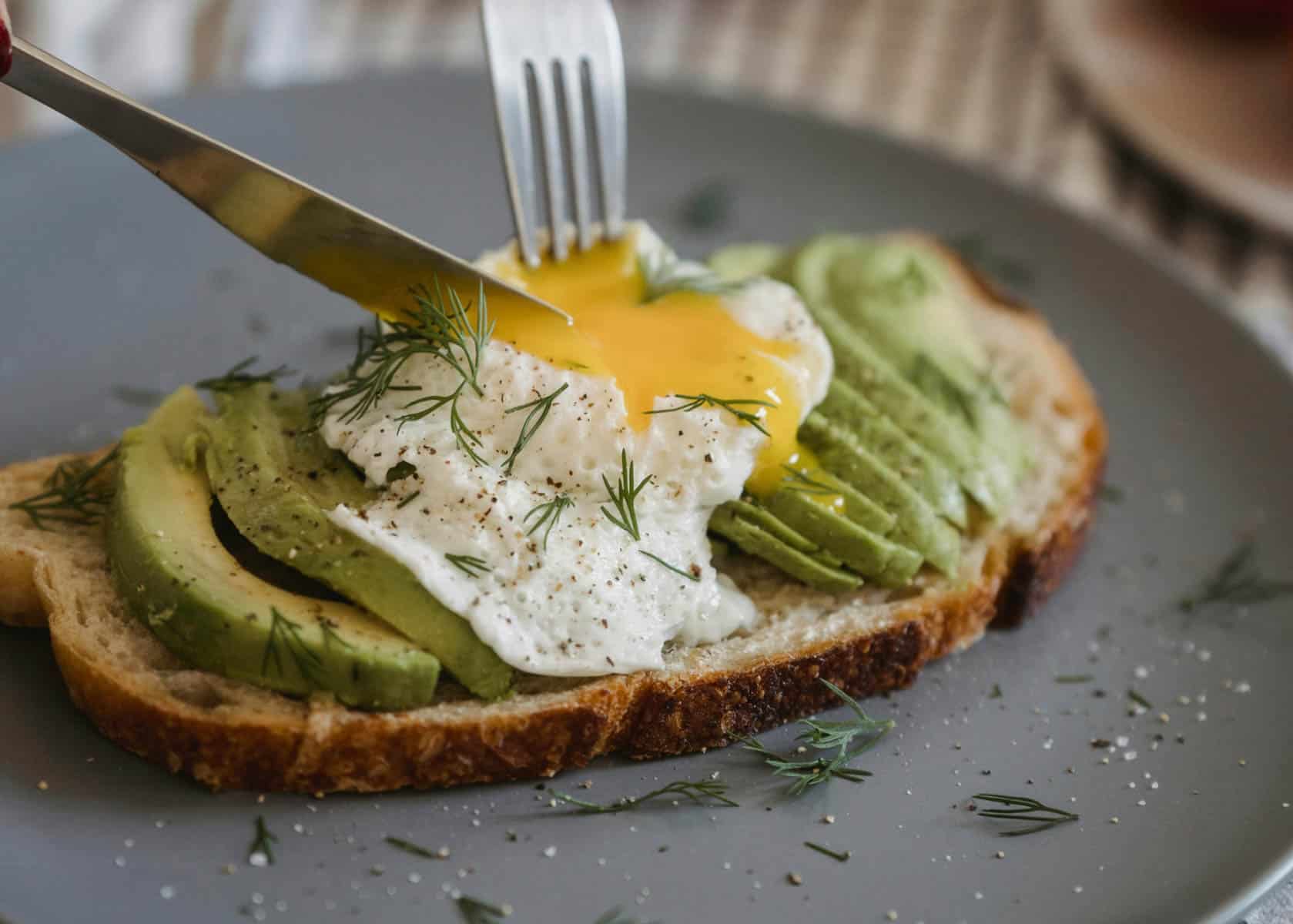 cutting poach egg on top of Sardine avocado toast on a plate with fresh cilantro and lemon wedges