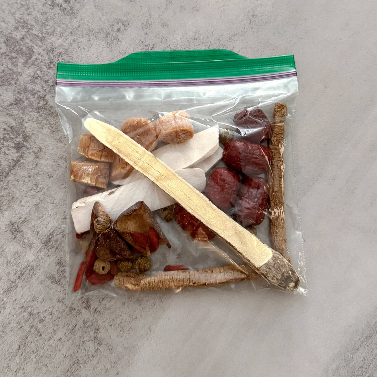 Clear resealable bag containing dried herbs, root slices and red dates for dendrobium silkie chicken soup on a grey surface.