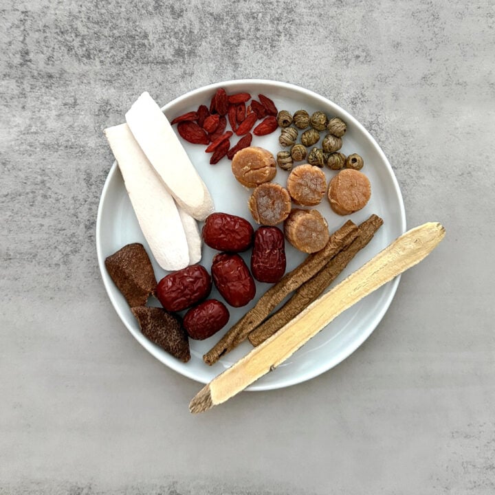 Dried herbs, root slices and red dates arranged on a white plate with surrounding grey backdrop, ready for silkie chicken soup.